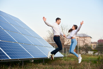 Young family jumping holding hands near the house with solar panels. Guy and girl are dressed in jeans, shirts and white sneakers are looking at each other. Solar energy concept image - Powered by Adobe