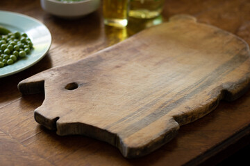 wooden cutting board shaped like a piggy, a plate with green peas and a bottle of oil on a wooden table.