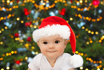 winter holidays, childhood and people concept - cute baby girl in santa hat over christmas tree lights on background