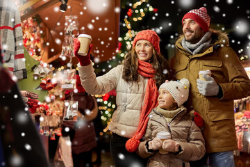 Fototapeta na wymiar family, winter holidays and celebration concept - happy mother, father and little daughter with takeaway drinks at christmas market on town hall square in tallinn, estonia over snow