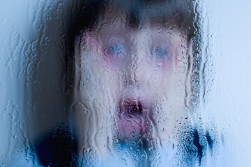 Children's fear and anxiety, psychological violence and pain. Young girl cries and screaming behind wet glass. Selective focus.