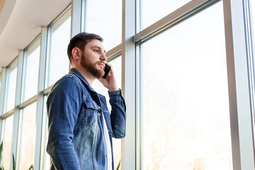 Young man making business call talking by phone indoors near window wall in casual jean jacket. Smart man in modern city office have mobile conversation. Caucasian bearded business man.