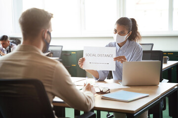 Business as new normal. Young mixed race woman wearing protective face mask showing paper with text SOCIAL DISTANCE to male colleague while working together in office