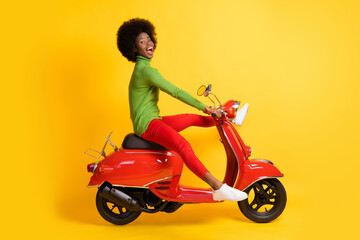 Obraz na płótnie Canvas Photo portrait of excited african american woman driving a red scooter with legs spread wearing casual outfit isolated on vivid yellow colored background
