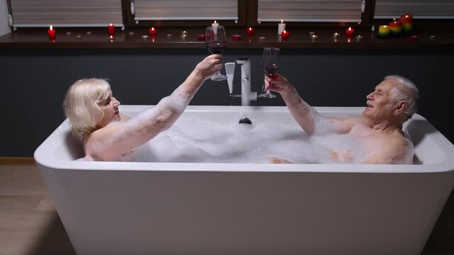 Sexy senior couple grandfather and grandmother is taking foamy bath, drinking red wine in luxury bathroom with candles. Elderly man and woman in love. Spa procedures in hotel. Honeymoon. 6k downscale