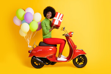Photo portrait of brunette african american girl holding red wrapped gift riding bike with air balloons isolated on vivid yellow colored background