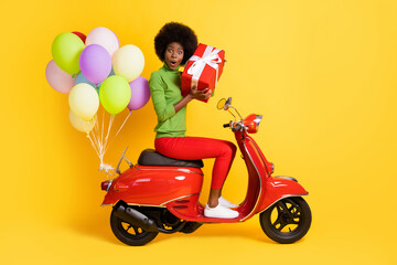 Photo portrait of african american girl holding box guessing what's inside wearing casual green pullover on red bike with air balloons isolated on vivid yellow colored background