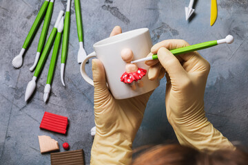 The master sculpts a human figurine on a white ceramic mug made of polymer clay, top view
