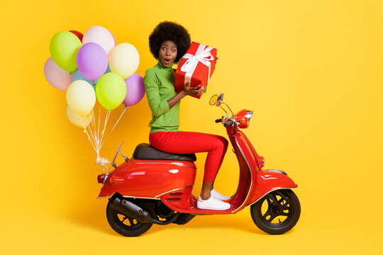 Photo portrait of african american girl holding box guessing what's inside wearing casual green pullover on red bike with air balloons isolated on vivid yellow colored background