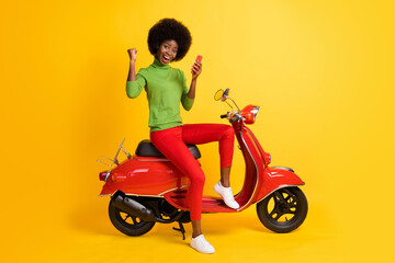 Obraz na płótnie Canvas Photo portrait of young brunette african american biker on red bike holding cellphone receiving winning sms message wearing casual outfit isolated on vivid yellow colored background