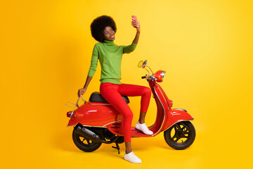 Fototapeta na wymiar Photo portrait of young rider biker with brunette sitting on red retro motorbike holding cellphone taking selfie isolated on vivid yellow colored background