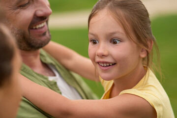 Close up of happy little girl looking aside while her dad holding her, spending time with family outdoors on a warm day