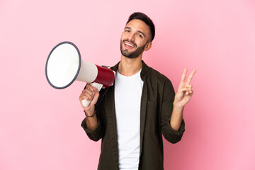 Young caucasian man isolated on pink background holding a megaphone and smiling and showing victory sign