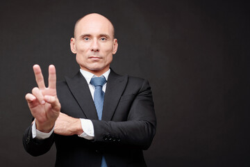 Portrait of serious mature bald politician in suit showing peace gesture and looking at camera