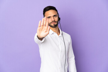 Telemarketer man working with a headset isolated on purple background making stop gesture