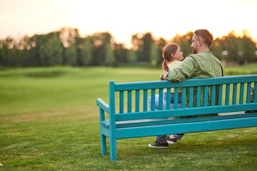 Full length shot of young father spending time with his cute little daughter. They are looking at each other while sitting on the bench in the park on a summer day