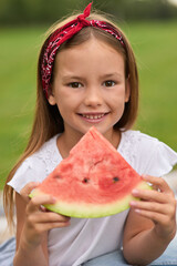 Portrait of cheerful little girl looking at camera while holding watermelon slice, family having a picnic in the green park on a summer day