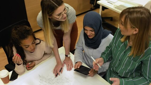Group of modern multicultural young women in casual wear discussing architectural designs in the creative office.