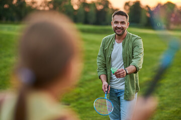 Happy father holding badminton racket and shuttlecock while playing with his little daughter outdoors in the park on a summer day