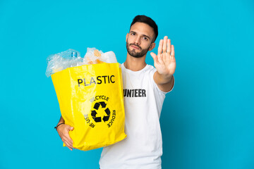 Young caucasian man holding a bag full of plastic bottles to recycle isolated on blue background...