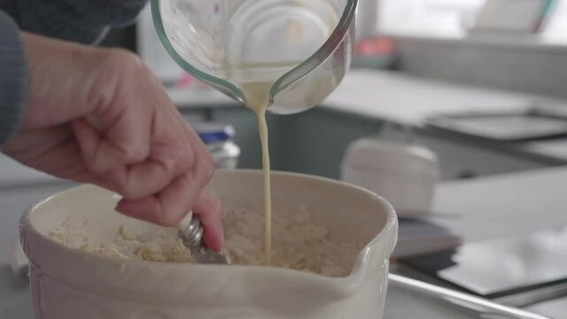 A lady making dough dough in the kitchen, she pours milk into the mixture in a ceramic bowl while stirring.