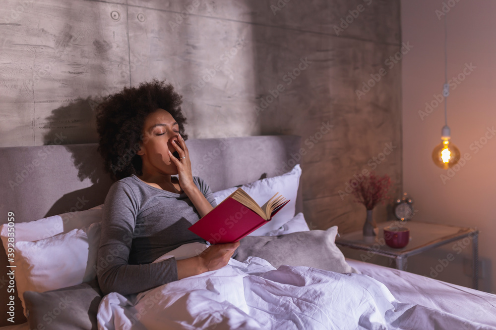 Wall mural Woman lying in bed yawning and reading a book