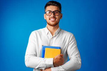 Man student in glasses holding books in arms against blue background