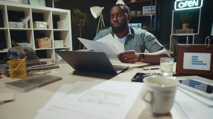 Fototapeta na wymiar Portrait of focused african man reading documents near laptop in night office.