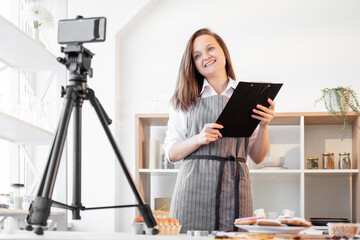 Online conference. Webinar training. Homemade culinary. Happy chef woman with black clipboard having videochat masterclass on smartphone light home kitchen interior.