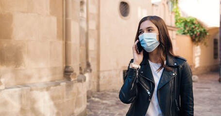 Pretty young Caucasian stylish woman in black leather jacket and medical mask strolling the street in town and talking on mobile phone. Beautiful female speaking on cellphone. Pandemic concept.