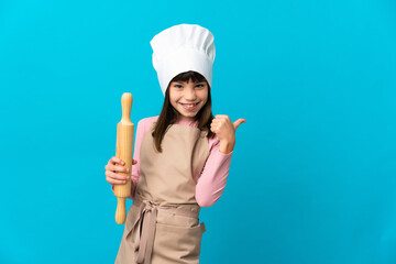 Little girl holding a rolling pin isolated on blue background pointing to the side to present a product