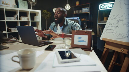 Emotional african man talking online in dark office. Afro guy looking camera