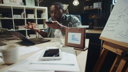 Fototapeta na wymiar Angry african hipster guy arguing on mobile phone in dark office.