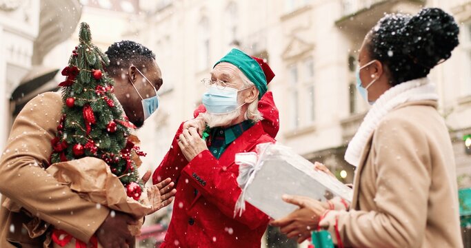 Portrait of happy African American man and woman with xmas presents standing outdoors and chatting while jld Caucasian Santa Clause in mask giving wrapped gift to female. Christmas miracle concept