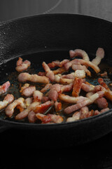 Chopped bacon lardons cooking on a hob in a cast iron frying pan