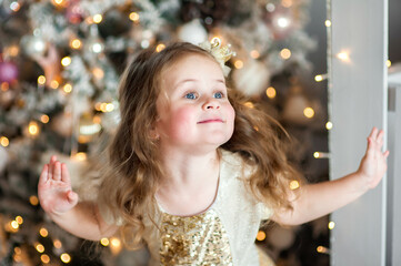 Portrait of a cute little girl with a crown on her head, making grimaces near the Christmas tree.