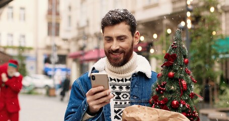 Handsome joyful Caucasian man standing in decorated crowded city and texting on smartphone and...