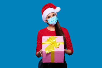 Fototapeta na wymiar Young woman wearing Santa hat and face protective medical mask holding christmas or new year gift box, blue isolated background.
