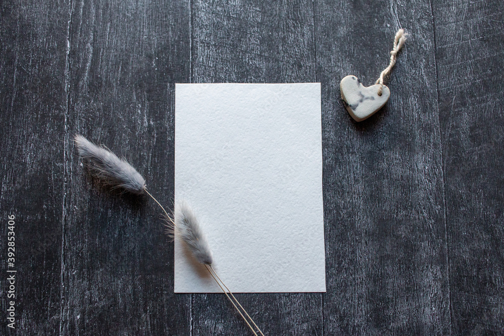 Wall mural Wedding invitation concept. White blank paper with  dry blue lagurus and ceramic heart shaped decoration on black wooden background