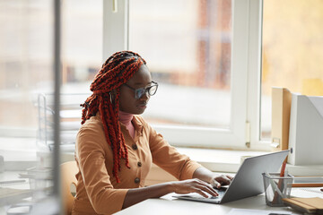 Portrait of young African-American businesswoman using laptop while sitting at desk by window and enjoying work in office, copy space