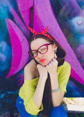 A stylish teenage girl in glasses and blue jeans poses near a graffiti wall