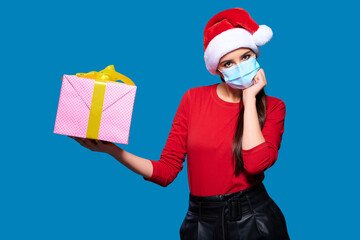 Upset woman wearing Santa hat and face protective medical mask and touching face, blue isolated background.