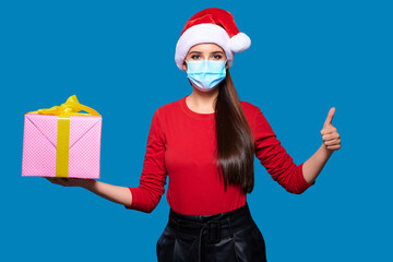 Fototapeta na wymiar Young woman wearing Santa hat wearing face protective medical mask holding christmas or new year gift box and showing thumb up gesture, blue isolated background.