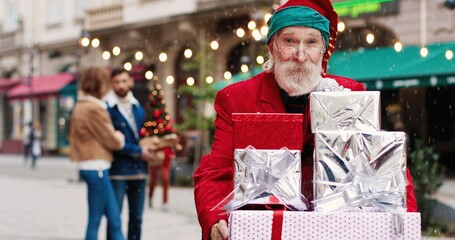 Portrait of happy senior Caucasian man in Santa Claus costume standing on crowded street and holding many christmas presents and kindly smiling. Holidays spirit. Xmas concept