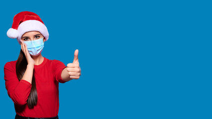 Fototapeta na wymiar Excited and shocked woman wearing Santa hat and face protective medical mask showing thumb up gesture, blue isolated background.