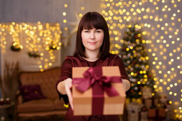 young beautiful woman giving Christmas present in decorated living room with Christmas tree and festive led lights