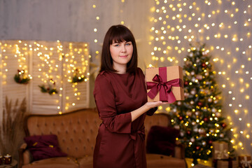 portrait of young beautiful woman with gift box in decorated living room with Christmas tree and festive led lights
