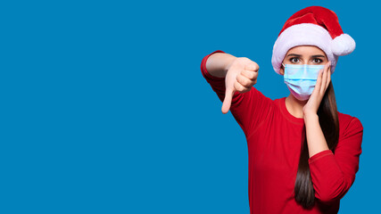 Fototapeta na wymiar Excited and shocked woman wearing Santa hat and face protective medical mask showing thumb down gesture, blue isolated background.