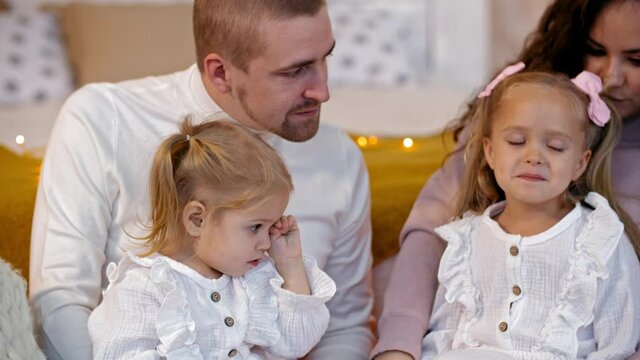 Hapyy Parent With Two Daughters Having Fun On Christmas Eve In Decorated Living Room. Cheerful Mom And Dad Amusing Sad Daughters On New Year Tree Background In Holiday Home