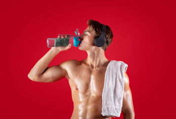 Sexy young sportsman with naked torso and towel listening to music in eadphones and drinking water...